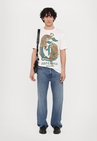 Biały t-shirt z grafiką w leopaarda, zestawiony z jasnoniebieskimi spodniami dzwonami i czarnymi butami. Czarna torba na ramię uzupełnia stylizację.