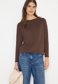 Bruine langemouwtop met een ronde hals, gecombineerd met blauwe denim jeans. De stof lijkt soepel met een ontspannen pasvorm. Een lichtbeige jas wordt in de hand gehouden.