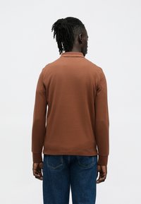 Bruine poloshirt met lange mouwen, een kraag, elastische manchetten en een gladde textuur. Draagt het met een blauwe spijkerbroek. Achteraanzicht toont ontwerpdetails.