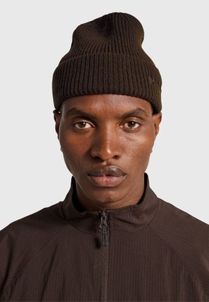 Brown knit beanie with a ribbed texture, featuring a slightly tapered crown. Worn with a matching dark zip-up jacket.