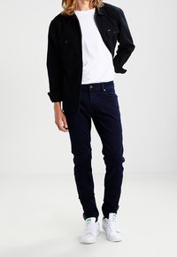 Chaqueta negra sobre una camiseta blanca, combinada con jeans azul oscuro y zapatillas blancas. El conjunto presenta un diseño casual y ajustado.