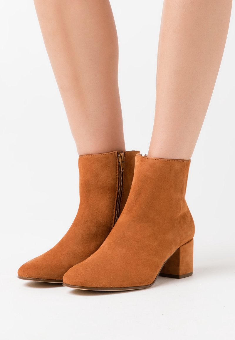 Bottes en daim couleur beige jusqu'à la cheville avec fermetures éclair latérales et talons blocs portées sur des jambes nues sur un fond blanc.