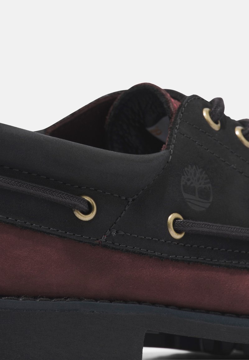 Zapato de ante negro y burdeos con puntera redonda, ojales dorados, cordones negros y logo estampado en el lateral. Suela de goma plana.