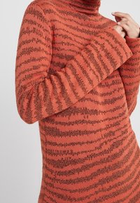 Femme portant un pull en tricot à col roulé rayé corail et noir à manches longues, les mains doucement jointes près du cou.