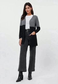 Šedý a černý kardigan s volnějším střihem, dlouhými rukávy a otevřeným předním dílem. Stylizován s bílým tričkem a černými džínami s vysokým pasem.