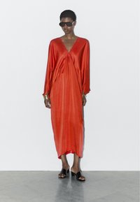 Luchtige jurk tot aan de enkels in levendig rood, met een diepe V-hals, brede mouwen en een gladde textuur, gecombineerd met zwarte sandalen en een zonnebril.