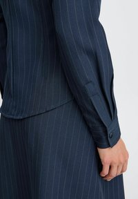 Camicia a maniche lunghe blu navy con righe, dotata di polsini con bottoni e vestibilità tailored. Il tessuto ha una texture liscia e una leggera lucentezza.