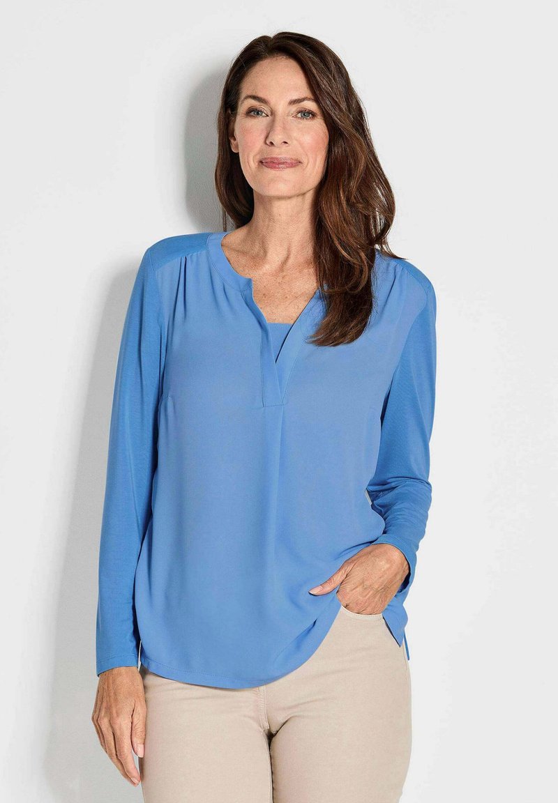 Himmelblaues Langarmbluse mit V-Ausschnitt, weicher Textur und abgerundetem Saum. Der Stoff liegt sanft am Körper an und hat dezente seitliche Schlitze.