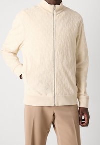 Man som bär en beige texturerad zip-up jacka och ljusbruna byxor, står med ena handen i fickan mot en enkel bakgrund.