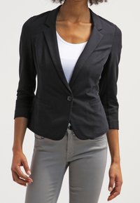 Musta bleiseri, jossa räätälöity istuvuus, 3/4-hiha, yksittäinen napitus ja etutaskut, yhdistettynä valkoiseen paitaan ja harmaihin housuihin.