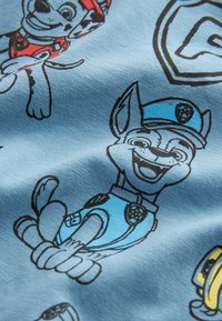 Vaaleansininen kangas, jossa on mustia viivapiirustuksia piirroskoirista eri unifor-mihoissa, punaisilla ja sinisillä yksityiskohdilla. Pehmeä, sileä pinta.