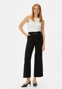 Zwarte wijdvallende broek met een gladde textuur en een metalen gesp detail op de tailleband, gecombineerd met een witte mouwloze top en zwarte hakken.