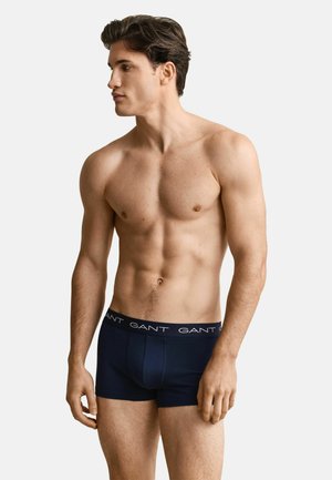 Jonge gespierde man kijkt naar links, draagt marineblauwe GANT boxershorts, staat tegen een effen witte achtergrond.