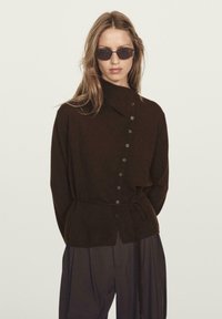 Camicia marrone con colletto alto e cintura legata, realizzata in tessuto morbido. Presenta bottoni laterali e una vestibilità rilassata con una trama sottile.