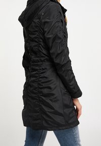 Manteau matelassé noir avec capuche, côtés froncés avec plissé, et texture lisse et brillante. Vue de dos montrant une silhouette ajustée et un détail de la capuche.