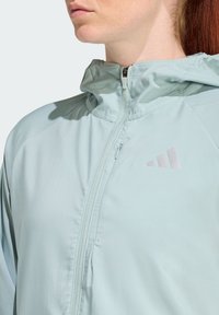 Svetle modrá zipová bunda s vysokým límcem, vyrobená z hladkého materiálu, s malým růžovým logem a jemnými detaily prošívání.