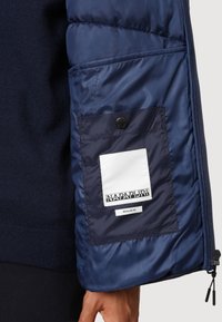 Manica e lato di giacca imbottita blu navy con tasca interna con bottone che mostra l'etichetta bianca "Napapijri" e il tag "Fascita regolare".