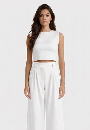 Donna con lunghi capelli scuri indossa un crop top bianco senza maniche e pantaloni bianchi a vita alta e gamba larga, in piedi davanti a uno sfondo chiaro semplice.