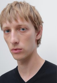 Giovane con capelli biondi corti e occhi blu, che indossa una maglietta nera a scollo a V su uno sfondo semplice e chiaro.