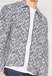 Tekstureret jakke med et marineblåt og hvidt plettet mønster, der har en lynlåslukning foran, kort krave og slank pasform.