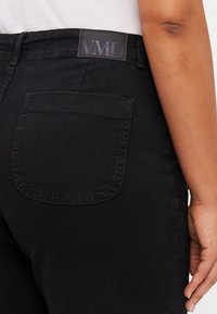 Svarta denimjeans med en slät yta, som har en bakficka och ett märkeslappar med texten "VMD" på midjebandet.