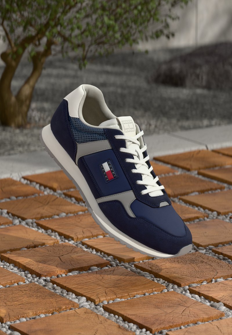 Marineblauwe sportschoen met een mix van suede en mesh, witte accenten, een grijze paneel en een logo-detail aan de zijkant.