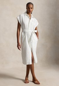 Woman wearing a white belted sleeveless midi shirt dress with front buttons and brown flat sandals against a neutral background.