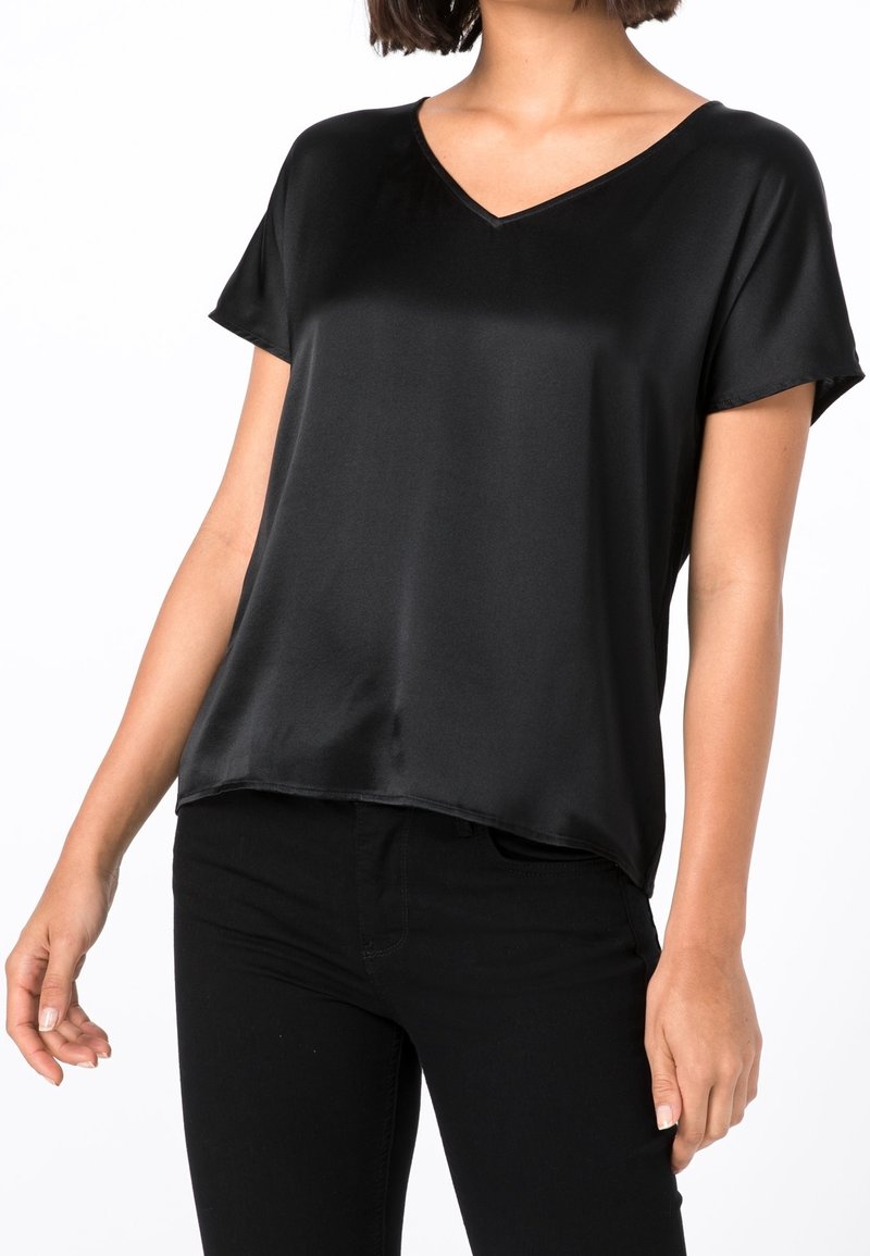 Femme portant un haut en satin noir à manches courtes et col en V, associé à un pantalon noir, vue de la moitié du torse jusqu’aux épaules sur fond blanc.