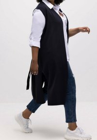 Chaleco largo negro de punto con lados abiertos, usado sobre una camisa blanca con cuello y combinado con jeans azules y zapatillas blancas.