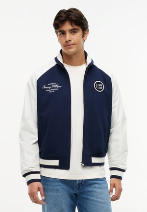 Jonge man die een marineblauw-wit varsityjack met geborduurde logo's draagt over een wit hemd en blauwe jeans, staand tegen een effen achtergrond.