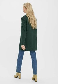 Dunkelgrüner, langer Cardigan mit lockerer Passform, kombiniert mit hellblauen Jeans und strukturierten goldenen High-Heels. Glatter Stoff, minimalistisches Design.