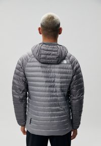 Jachetă puffer gri cu un design matlasat, glugă atașată și logo pe partea din spate superioară. Prezintă mâneci lungi și un tiv drept.