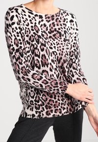 Kobieta w długim rękawie sweter w leopardzie wzór w czerni, beżu i różu, zestawiony z czarnymi spodniami, pozująca z rękami skrzyżowanymi.