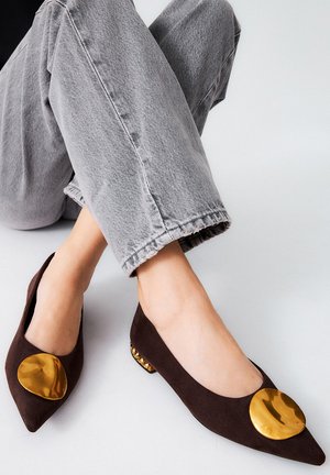 Zapatos planos de ante marrón con un gran acento metálico dorado en la puntera y un pequeño tacón con tachuelas doradas. Combinados con pantalones de mezclilla cortos grises.