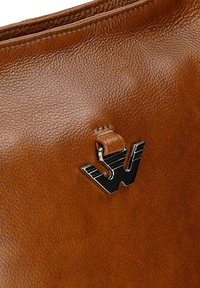 Brązowa skórzana powierzchnia z teksturowanym ziarnem oraz małą czarną metalową emblematem "W" przymocowaną za pomocą skórzanej pętli.