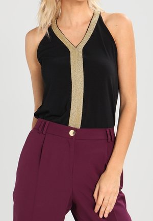 Femme portant un haut noir sans manches avec une bande dorée en forme de V et un pantalon prune taille haute avec un bouton doré, main posée sur la hanche.