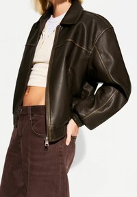 Braune Lederbomberjacke mit Reißverschluss, gerippten Bündchen und Kragen. Tragend über einem hellbeigen Oberteil und kombiniert mit dunkelbraunen Hosen.
