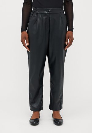 Person trägt schwarze, hoch taillierte, verjüngte Hosen aus Kunstleder, kombiniert mit einem schwarzen langärmligen Oberteil und schwarzen Ballerinas.