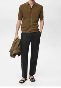 Camisa de punto marrón de manga corta con botones, pantalones negros ajustados, chaqueta verde colgada del brazo y sandalias negras en los pies.