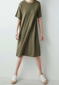 Olive green cotton jersey dress, short sleeves, round neckline with yellow trim, loose fit, falls above the knee, paired with white sneakers.