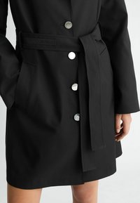 Cappotto lungo nero realizzato in tessuto liscio, con bottoni metallici, cintura in vita e tasche laterali. Il design è semplice e funzionale.