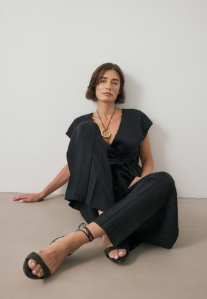 Jumpsuit negro con mangas cortas, textura plisada y cinturón en la cintura. Accesorios incluyen un collar con colgante dorado y sandalias planas negras.