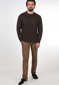 Brauner Strickpullover mit einem strukturierten Muster, kombiniert mit hellbraunen Slim-Fit-Hosen und schwarzen Sneakers mit weißen Sohlen.