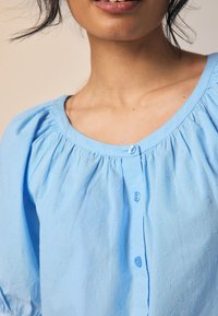 Hellblaue Bluse mit abgerundetem Ausschnitt, gerafften Details und kleinen, gestickten Mustern. Verfügt über Knöpfe an der Vorderseite und puffige Ärmel.