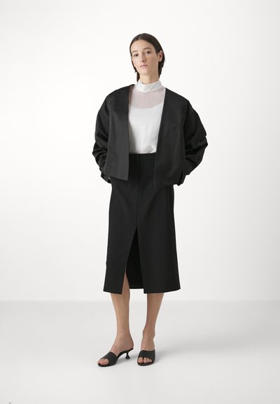 Black cropped bomber jacket over a white sheer top and black midi skirt with a front slit. Model wears black open-toe mules. Plain background.