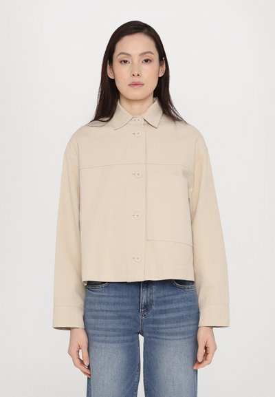 Beige kurz geschnittene Jacke aus Baumwollmischung, mit einer Knopfleiste, Umlegekragen und einer einzelnen Brusttasche. Getragen mit blauen Jeans.