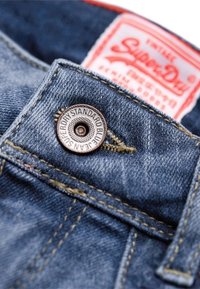 Džínsy z denimu s modrou texturou látky, s kovovým knoflíkem s vyraženým názvem značky a červenou látkovou etiketou na pásku.