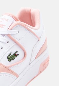 Λευκό και ροζ χαμηλό αθλητικό παπούτσι με πράσινο λογότυπο κροκόδειλου, λευκά κορδόνια, επενδεδυμένο κολάρο και υφή σόλας με το όνομα της μάρκας στη φτέρνα.