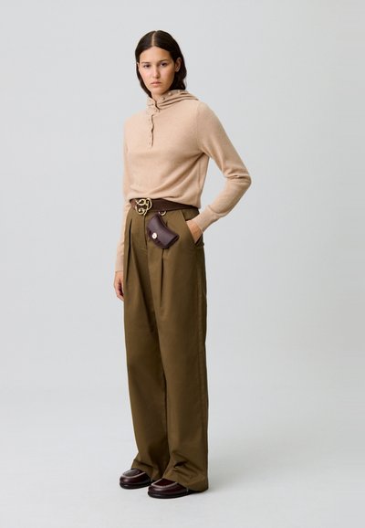 Pull en tricot beige avec capuche et patte de boutonnage, associé à un pantalon large vert olive et des shoes marron foncé. Petit sac marron accroché à la ceinture.