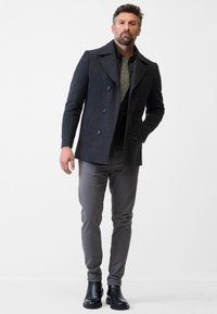 Donkergrijs wollen peacoat met grote knopen, gedragen over een lichtgrijze trui. Slim-fit grijze broek en zwarte enkellaarzen maken de look compleet.